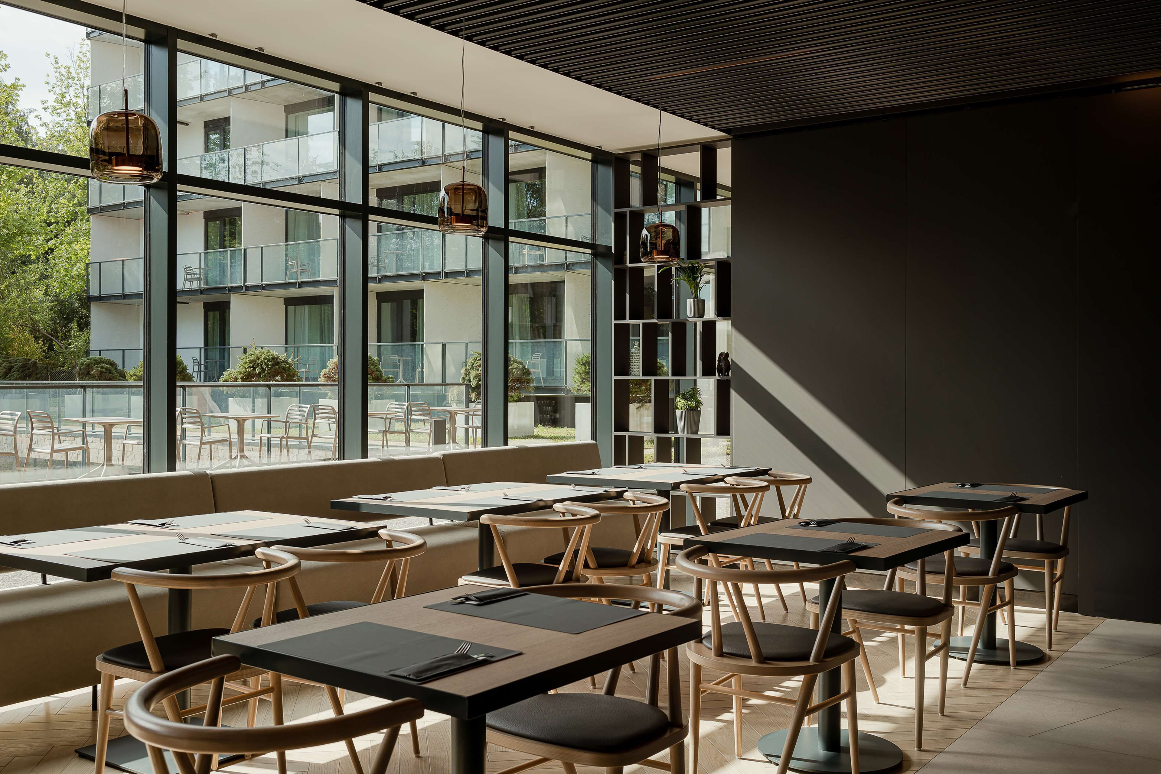 A modern restaurant with large windows, wooden furniture, and a view of a building with balconies.