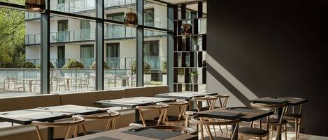 A modern restaurant with large windows, wooden furniture, and a view of a building with balconies.