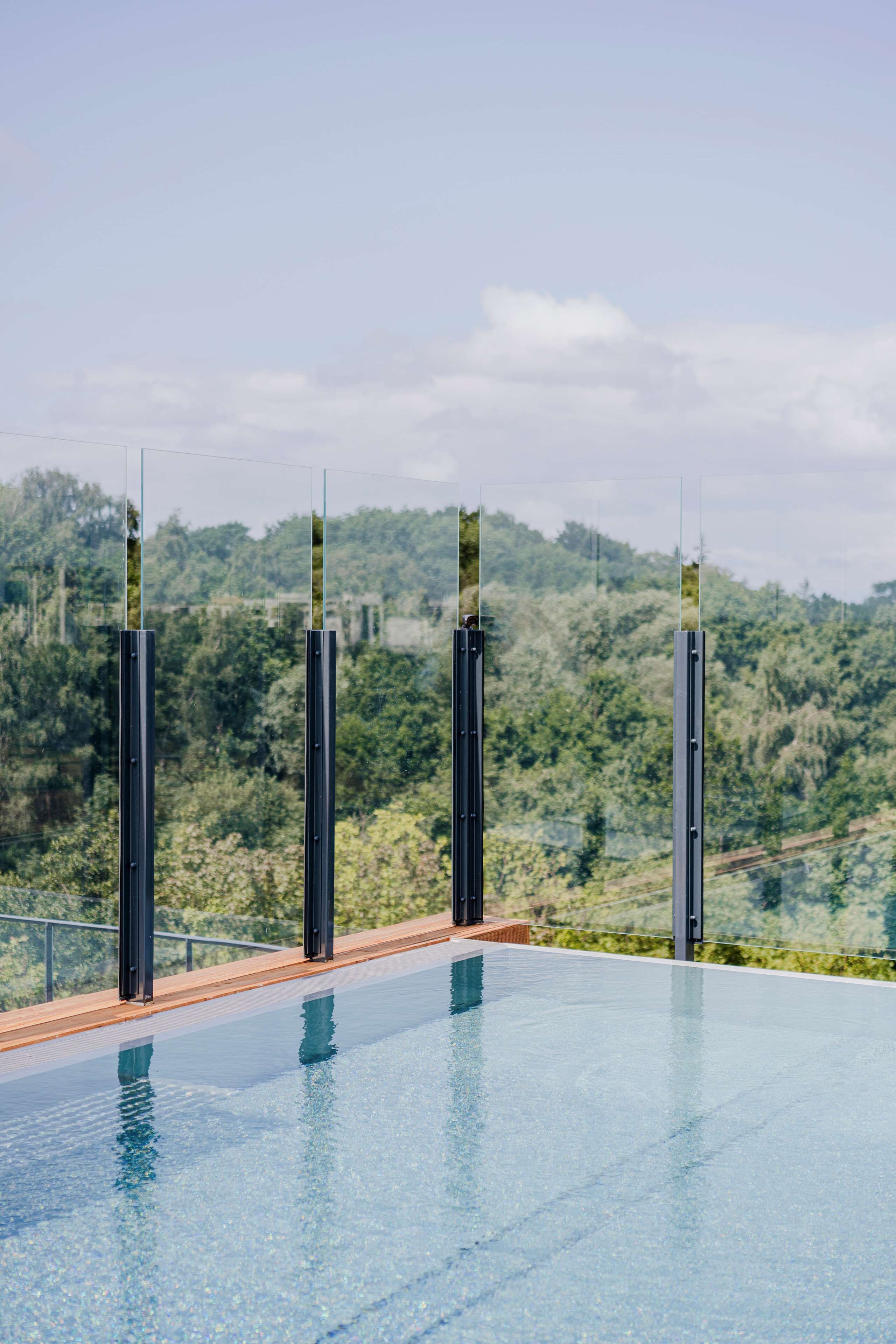 Una piscina infinita moderna con agua cristalina, rodeada de barandillas metálicas y una terraza de madera, con vistas a una exuberante ladera verde.