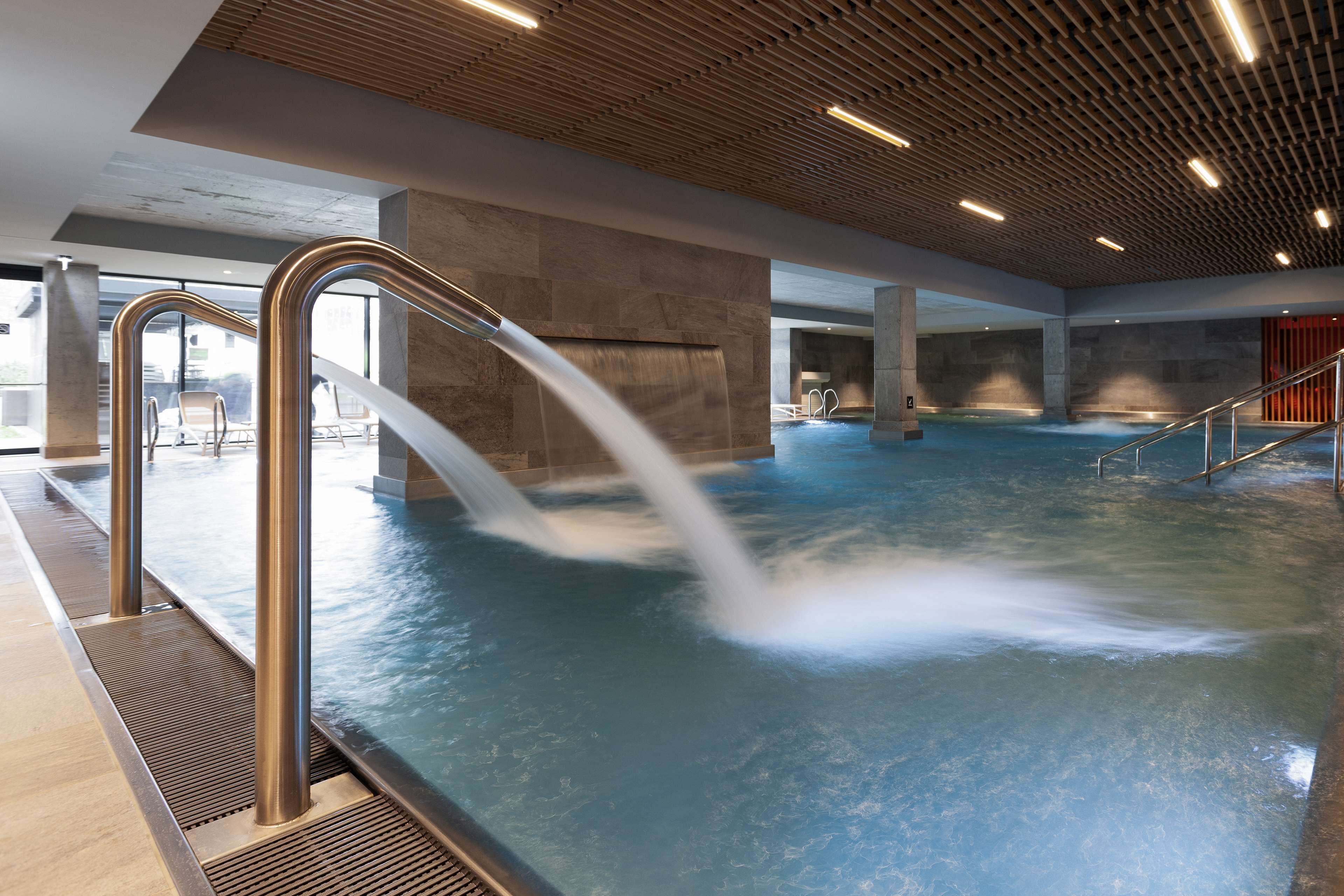 Una amplia piscina interior con múltiples chorros de agua, techo de madera y diseño moderno.