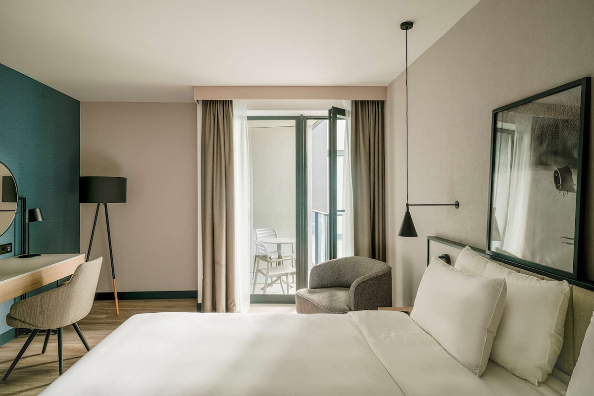 A hotel room with a bed, a desk, a chair, a lamp, and a large window with curtains.