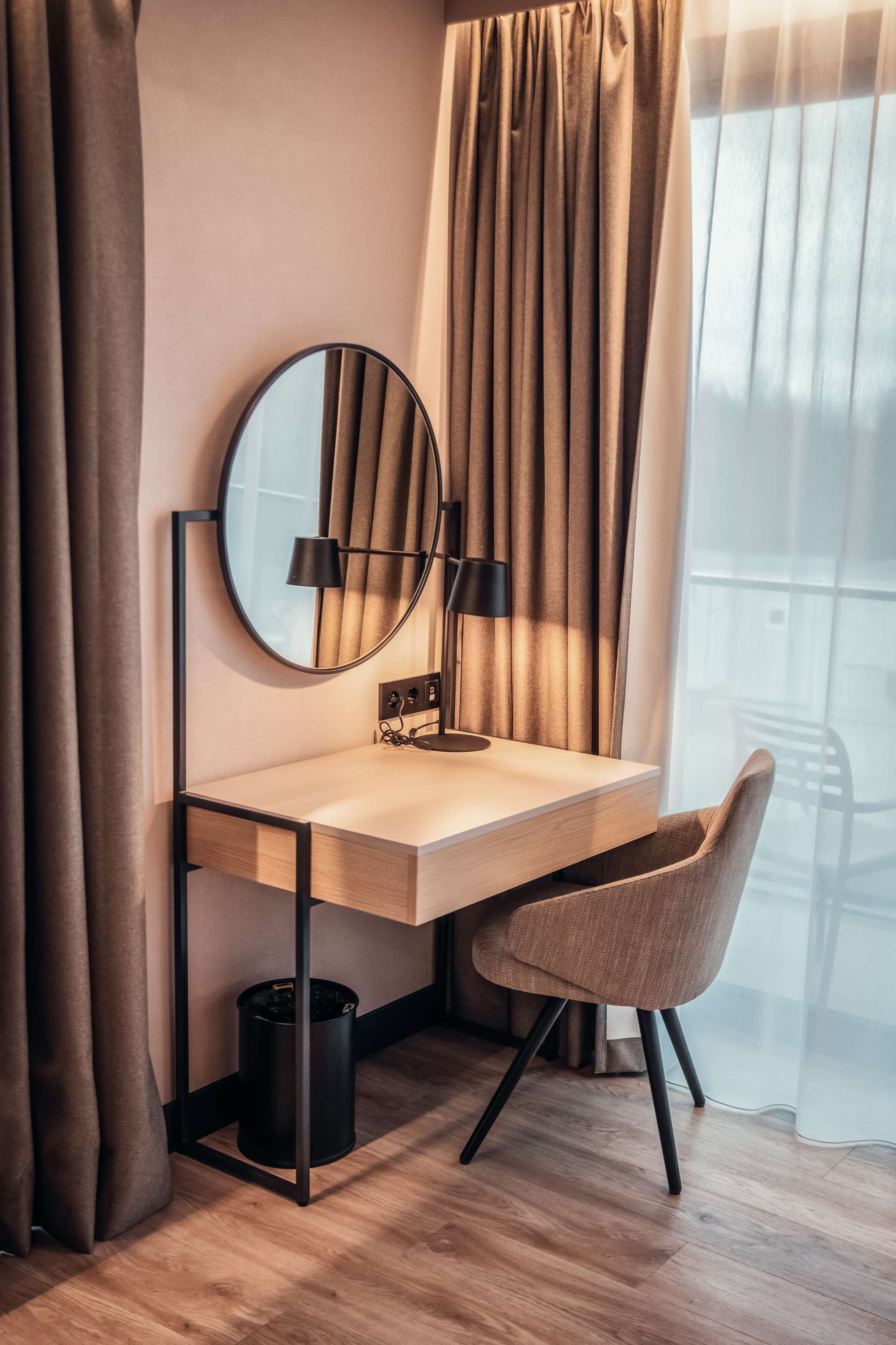 A modern hotel room with a wooden desk, a round mirror, a black lamp, and a chair.
