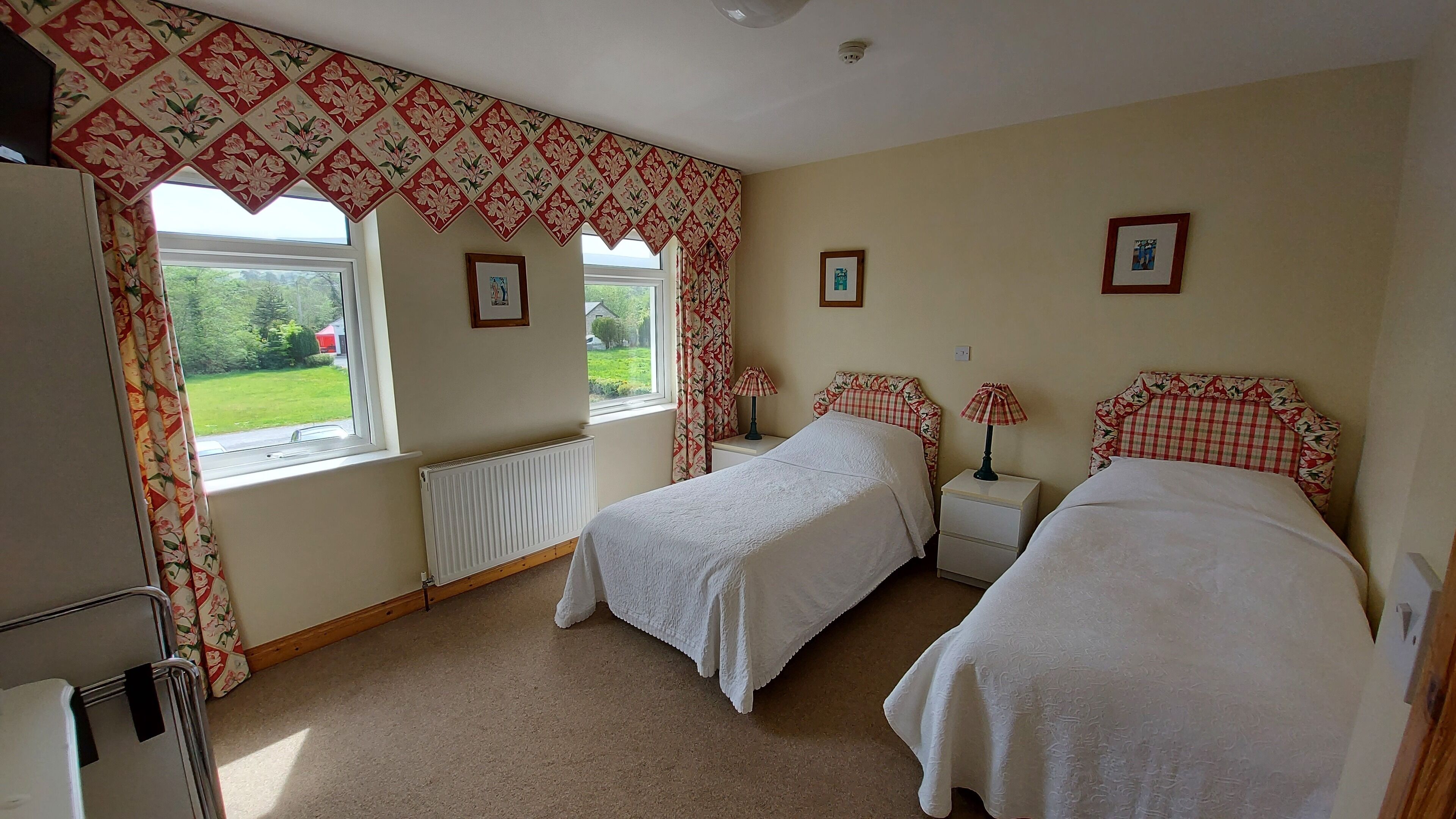 A bedroom with two beds, a radiator, a window with curtains, and a lamp on a bedside table.