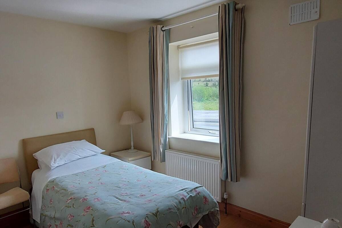 Een slaapkamer met een bed, nachtkastje, bureau, stoel, radiator, raam met gordijnen en een plafondlamp.
