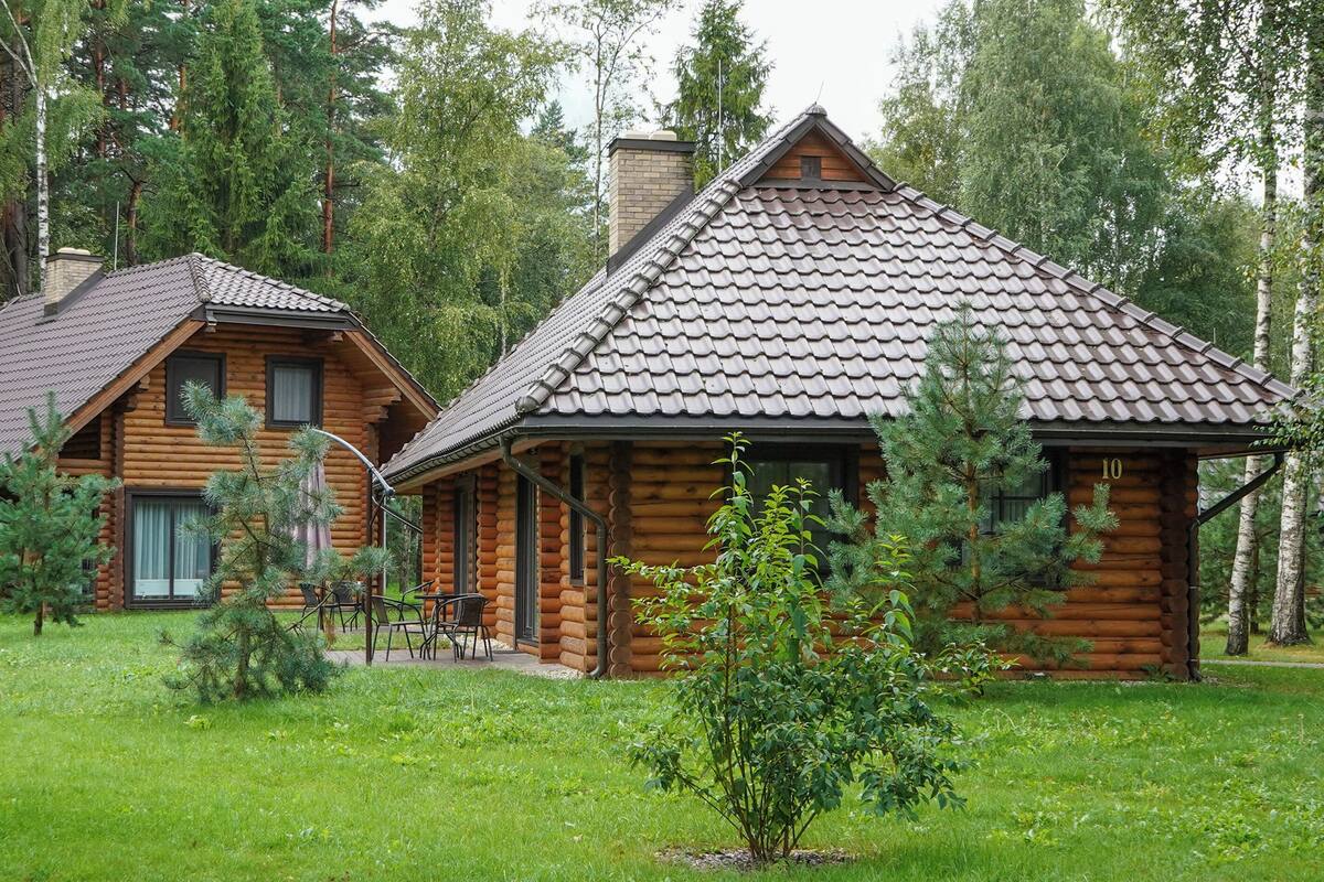 Två timmerstugor med sadeltak, omgivna av en skog. Varje stuga har en liten veranda med stolar.