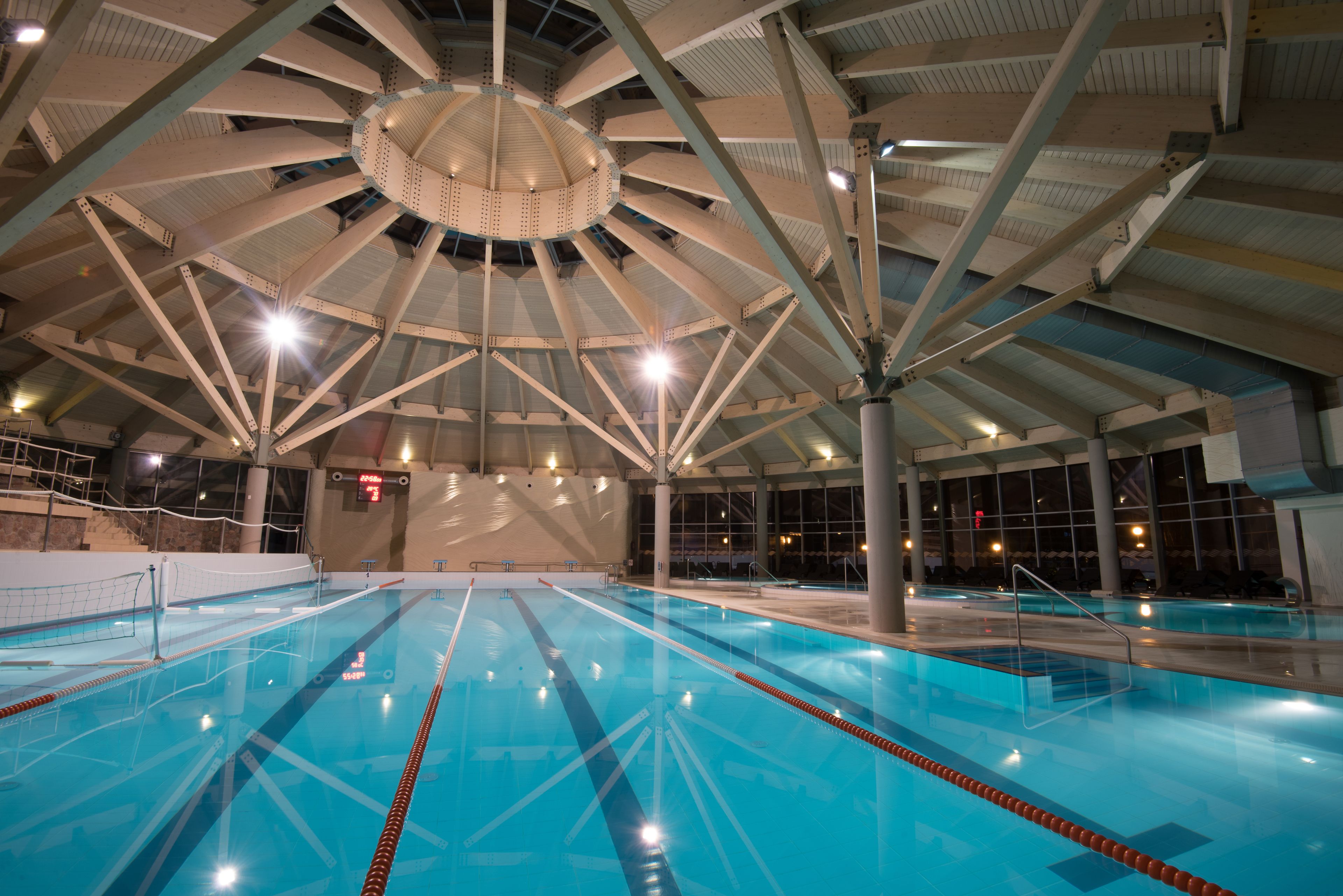 Une piscine intérieure avec des couloirs bien délimités, un plafond haut avec des poutres apparentes et de grandes fenêtres.