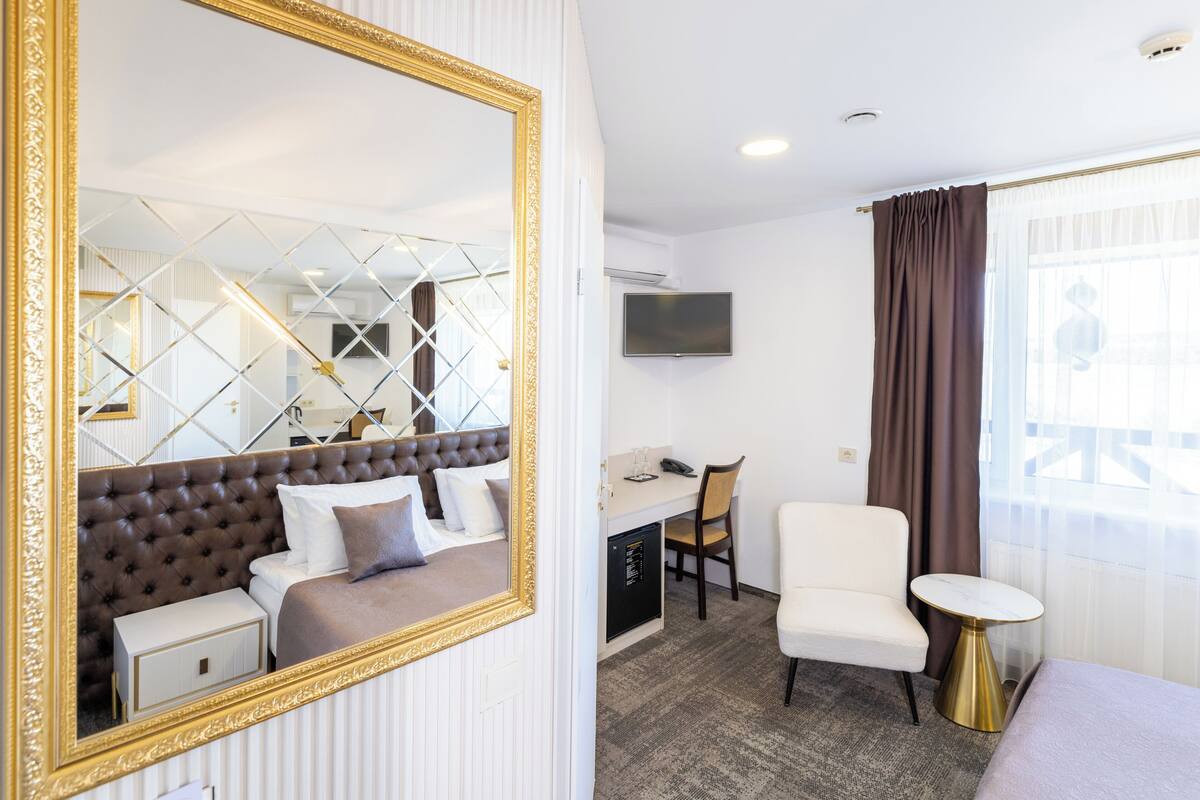 Ett hotellrum med en stor spegel, en säng med ett stoppat sänggavel, ett skrivbord med en stol, ett litet bord och en tv som är monterad på väggen.