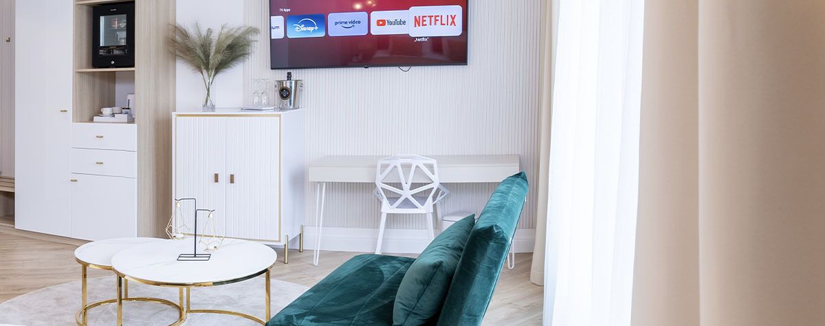 A modern living room with a flat-screen TV displaying streaming services, a white desk, a green sofa, and a white cabinet with drawers.