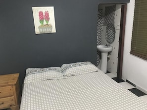 A bedroom with a bed, a wooden nightstand, a wall-mounted sink, and a door leading to another room. - Aarony House (Santo Domingo)