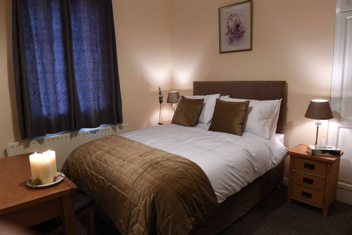 Una cama bien hecha con un edredón marrón, una mesita de noche con lámpara y una ventana con cortinas.