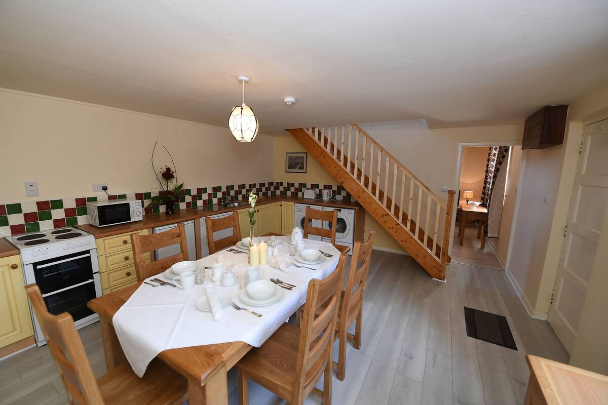 Una cocina con una mesa de comedor preparada para cuatro personas, sillas de madera, un microondas, una estufa y una lavadora.