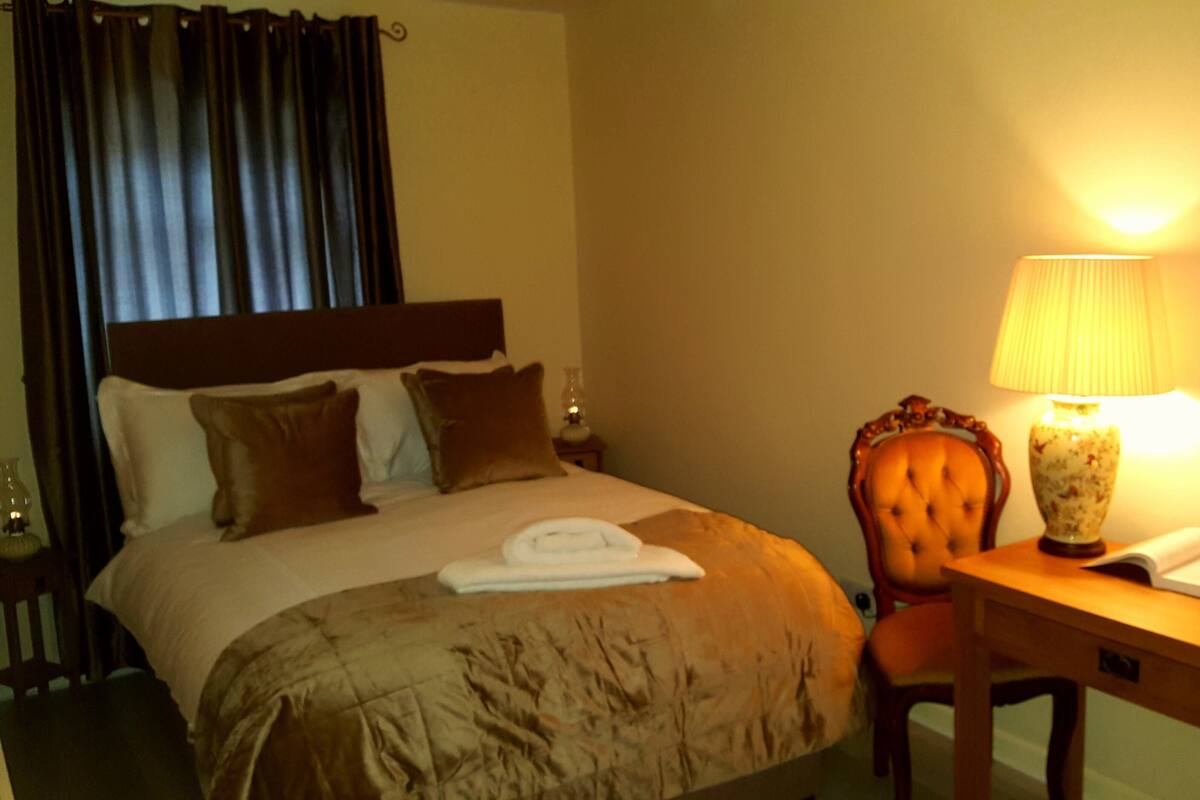 Habitación de hotel con cama, una silla, escritorio, lámpara y mesita de noche.