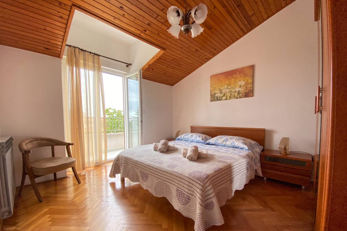 Un dormitorio con techo de madera, una cama grande con colcha estampada, una mesita de noche con lámpara, una silla y un ventanal corredero a un balcón.
