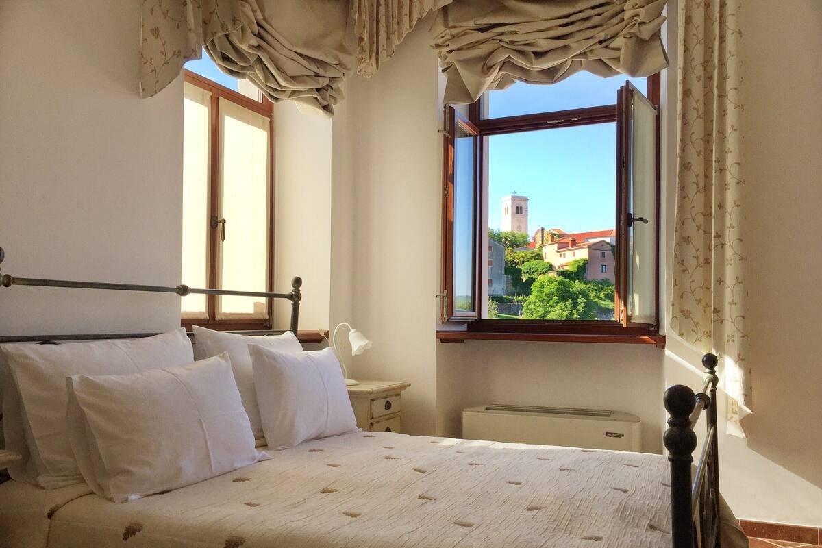 Une chambre à coucher comprenant un lit, une table de chevet, une lampe et une fenêtre donnant sur des immeubles et une tour.