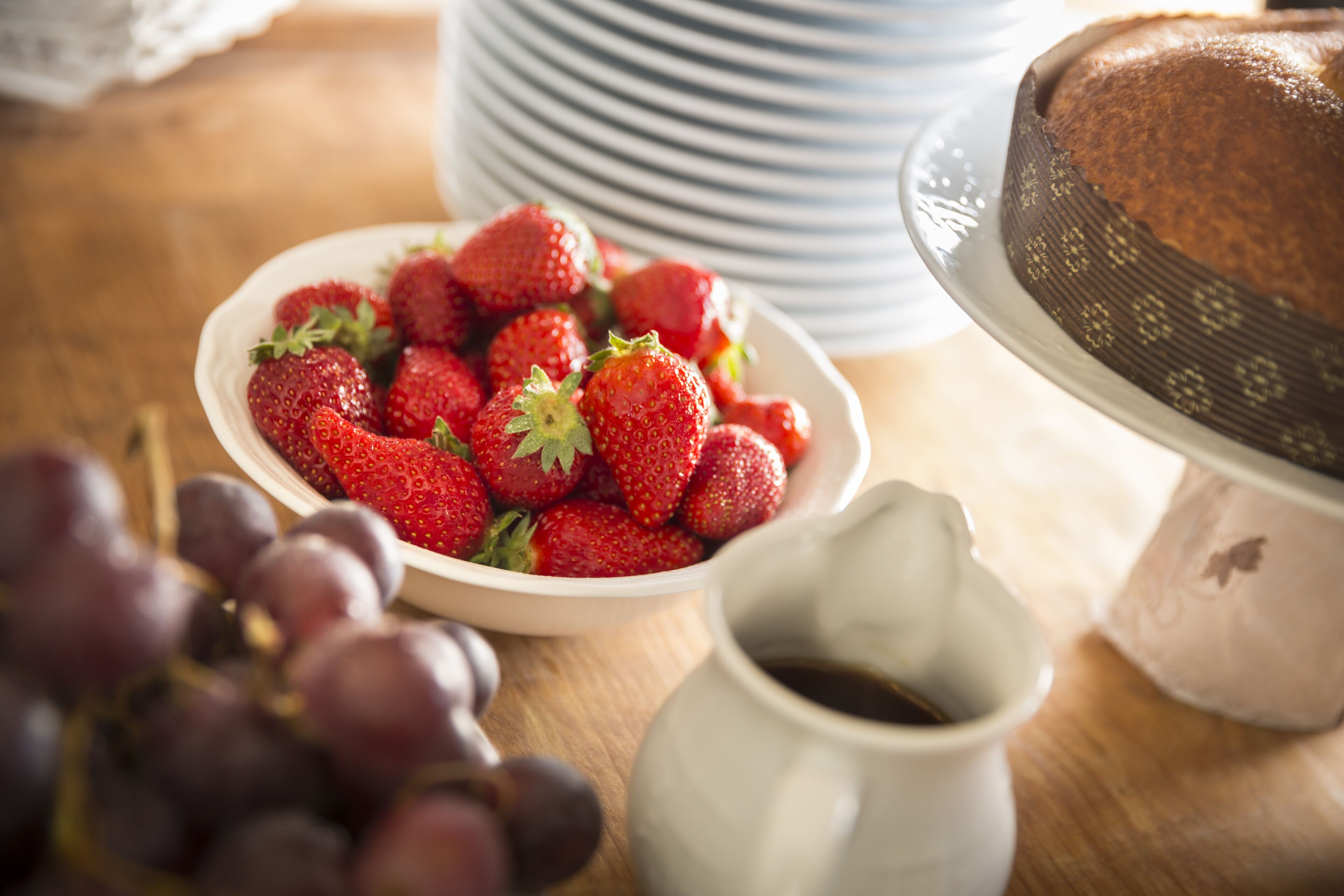 A bowl of strawberries, a cake on a stand, and a pitcher of coffee on a wooden table.