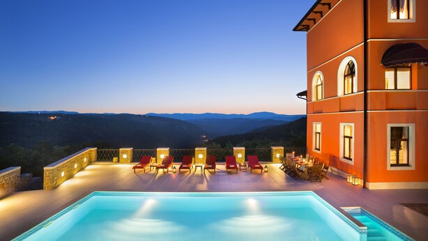 Ein Poolbereich mit Bergblick, Sitzgelegenheiten im Freien und ein Gebäude mit Rundbogenfenstern.