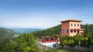 Ein Poolbereich mit roten Liegestühlen und Sonnenschirmen, umgeben von Steinmauern und Grünpflanzen, mit Blick auf eine Berglandschaft.
