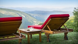 Zwei Holzliegen mit roten Kissen, ein kleiner Tisch mit einem Glas Wasser und ein malerischer Blick auf Berge im Hintergrund.