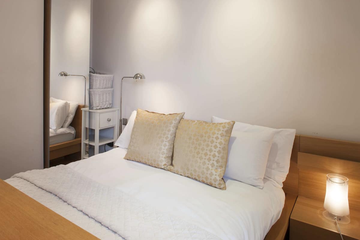 A neatly made bed with white bedding and two patterned pillows, a bedside table with a lamp, and a small white shelf with baskets.