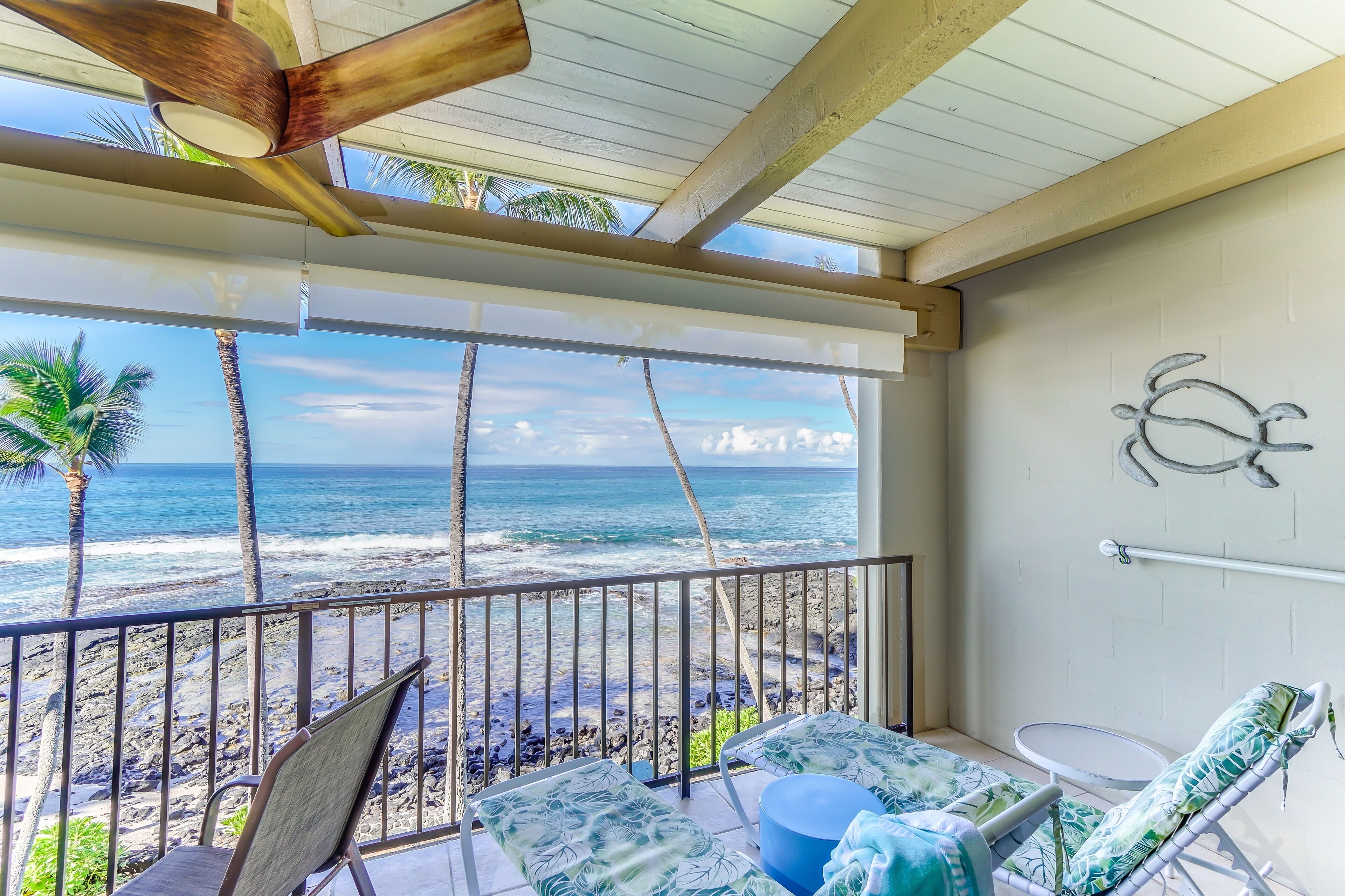 A balcony with a view of the ocean, featuring a ceiling fan, a railing, and lounge chairs.