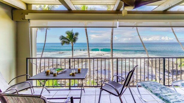 A balcony with a table and chairs, overlooking a beach with palm trees and the ocean.