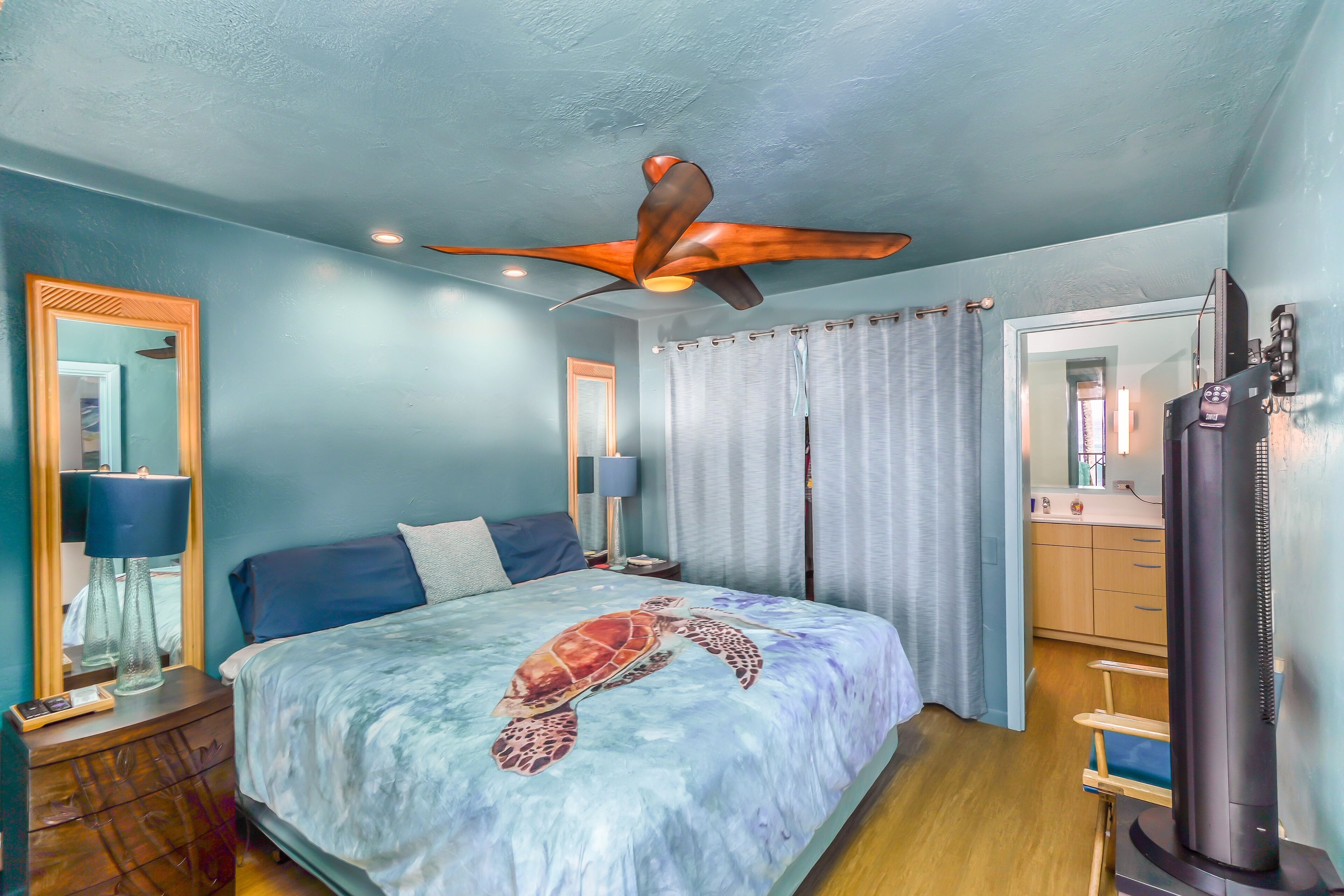A bedroom with a bed, a ceiling fan, a nightstand, a lamp, a mirror, a window with curtains, and a heater.