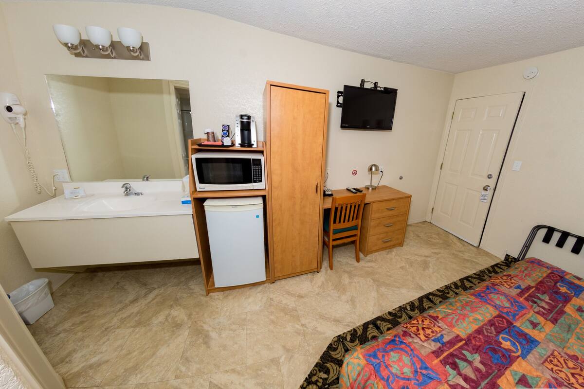 A bedroom with a bed, a desk, a chair, a microwave, a small fridge, a sink, a mirror, and a television.