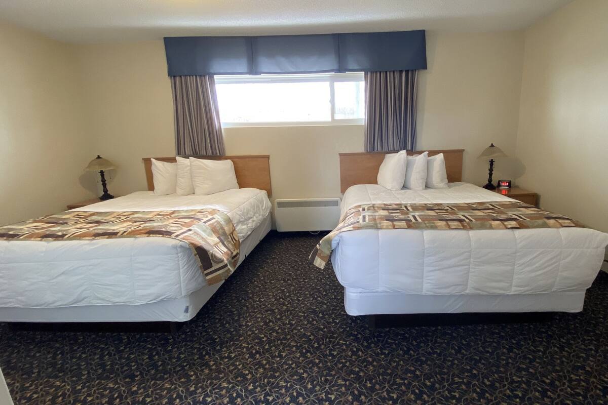A hotel room with two beds, a window with curtains, a heater, and a clock on the nightstand.