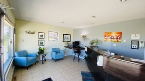 Un espace d’accueil avec un bureau en bois, deux chaises bleues, une petite table avec une plante, des tableaux encadrés au mur et une fenêtre donnant sur un paysage.