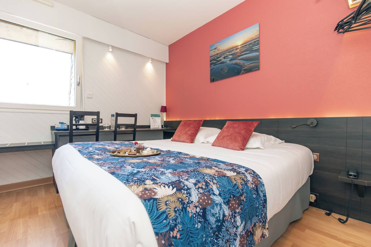 Une chambre d’hôtel avec un lit, un bureau, une chaise, un tableau au mur et une fenêtre.