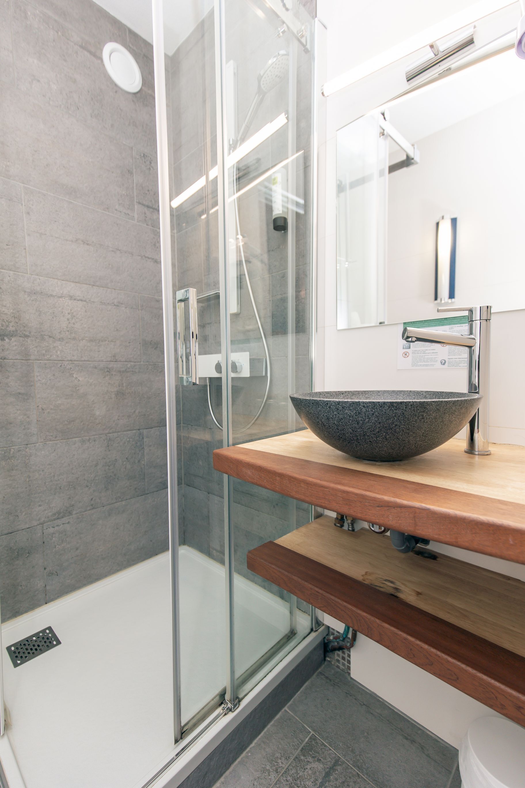 Un baño moderno con cabina de ducha de vidrio, tocador de madera con lavamanos de piedra y un espejo sobre el lavamanos.