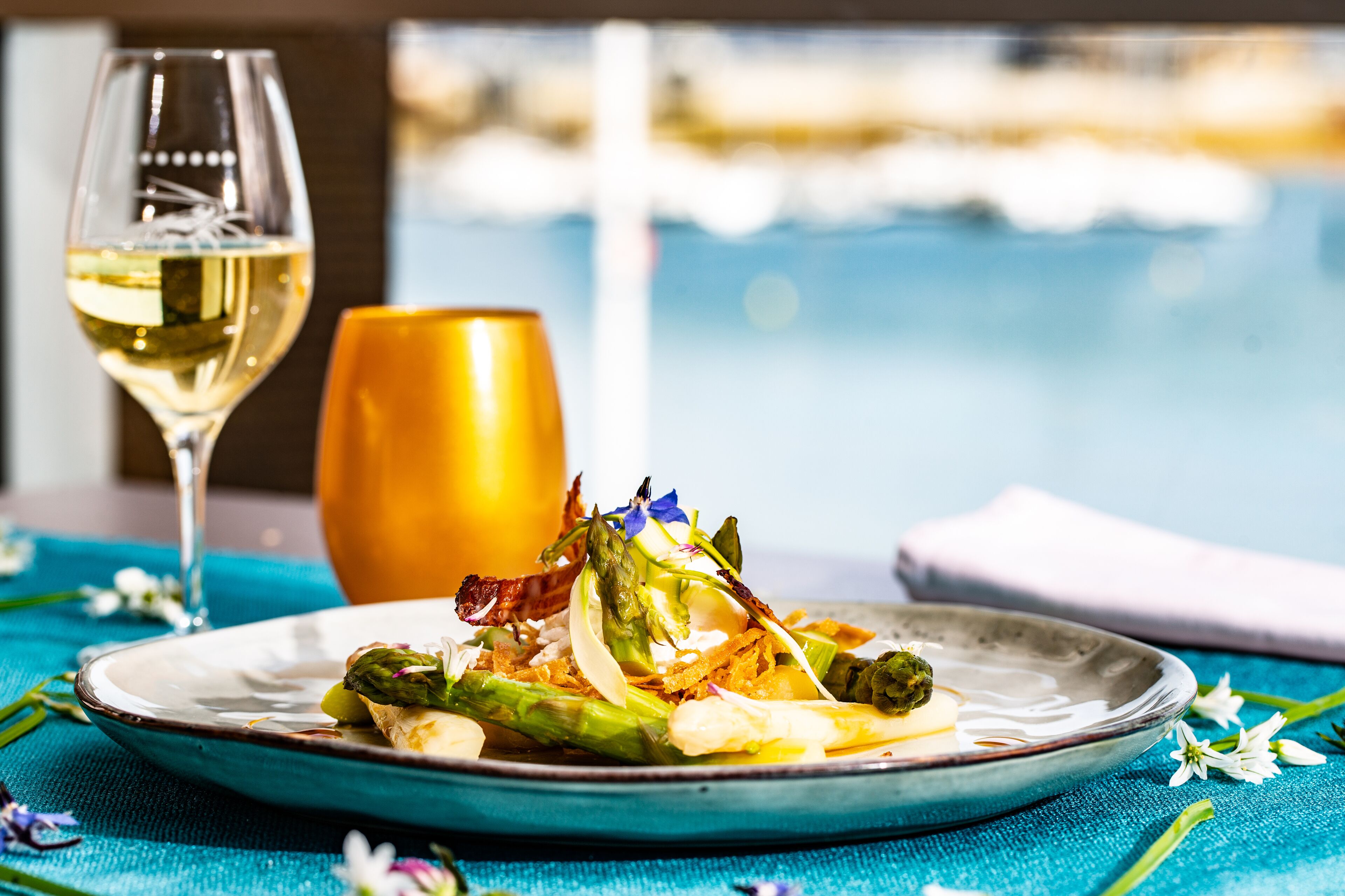 A plate of food with asparagus and other vegetables, a glass of white wine, and a golden vase on a table with a blue tablecloth.