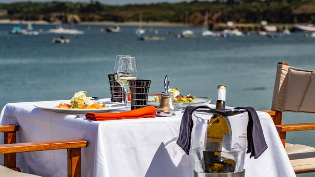 A table set for a meal with a bottle of wine, a glass, and a pitcher, overlooking a body of water with boats. - Logis Hôtel de France - Face à la Mer (Camaret-sur-Mer)