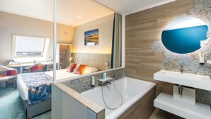 A modern hotel room with a large bed, a sitting area with a view, a bathroom with a large mirror and sink. - Logis Hôtel de France - Face à la Mer (Camaret-sur-Mer)