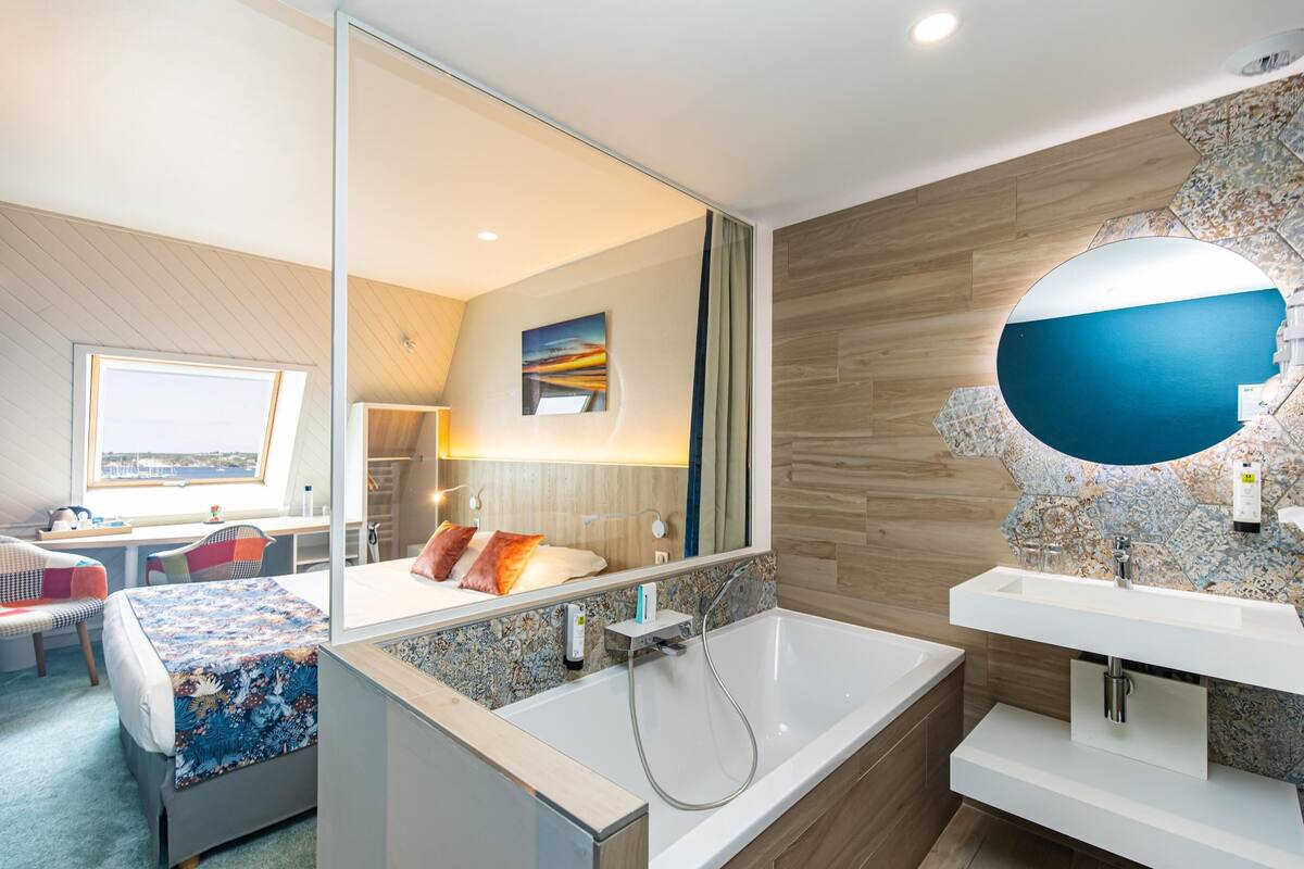 Une chambre d’hôtel moderne dotée d’un grand lit, d’un coin salon avec vue, et d’une salle de bain avec un grand miroir et un lavabo.