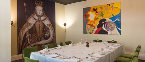 Un coin repas avec une longue table dressée pour six personnes, des chaises à l’assise verte et un grand tableau accroché au mur.