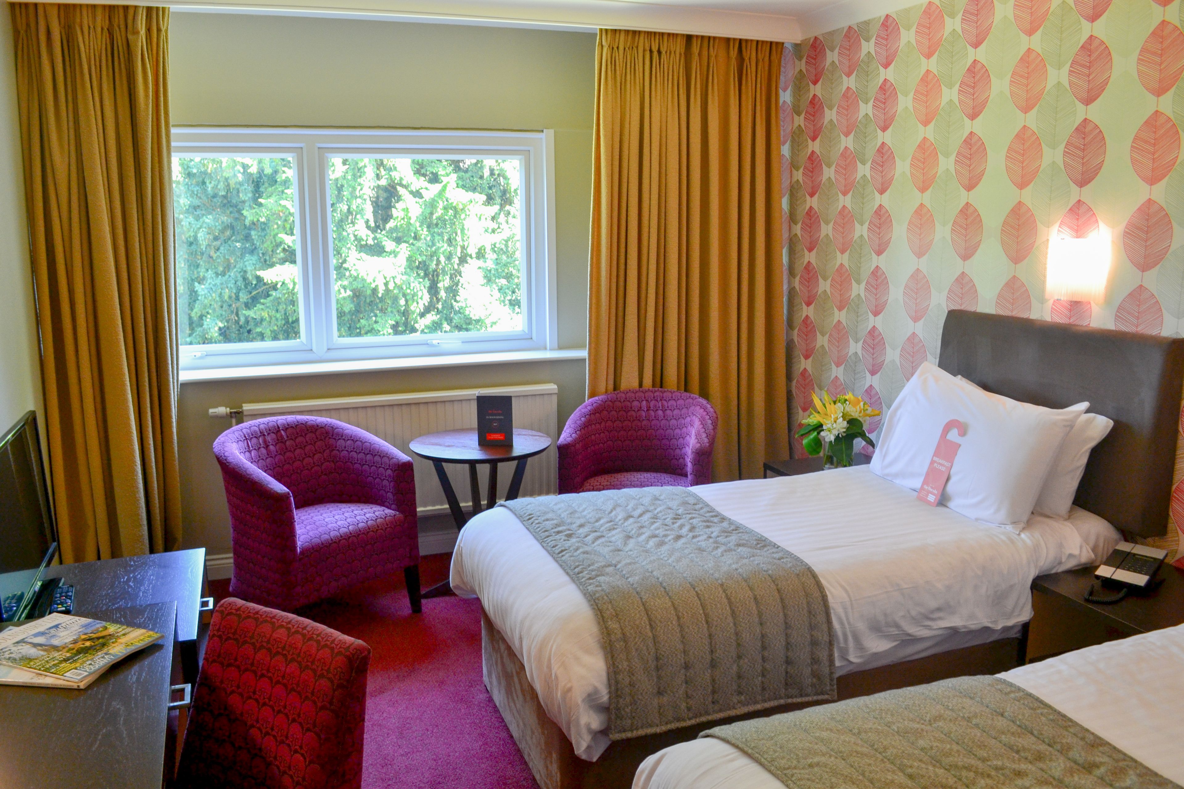 Ein Hotelzimmer mit einem Bett, zwei Sesseln, einem kleinen Tisch und einem Fenster mit Vorhängen.