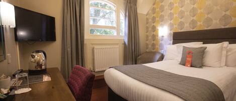 A hotel room with a bed, a desk with a kettle, a television, and a window with curtains.
