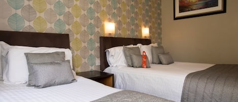 A hotel room with two beds, a patterned wallpaper, a framed painting, and a nightstand with a lamp.