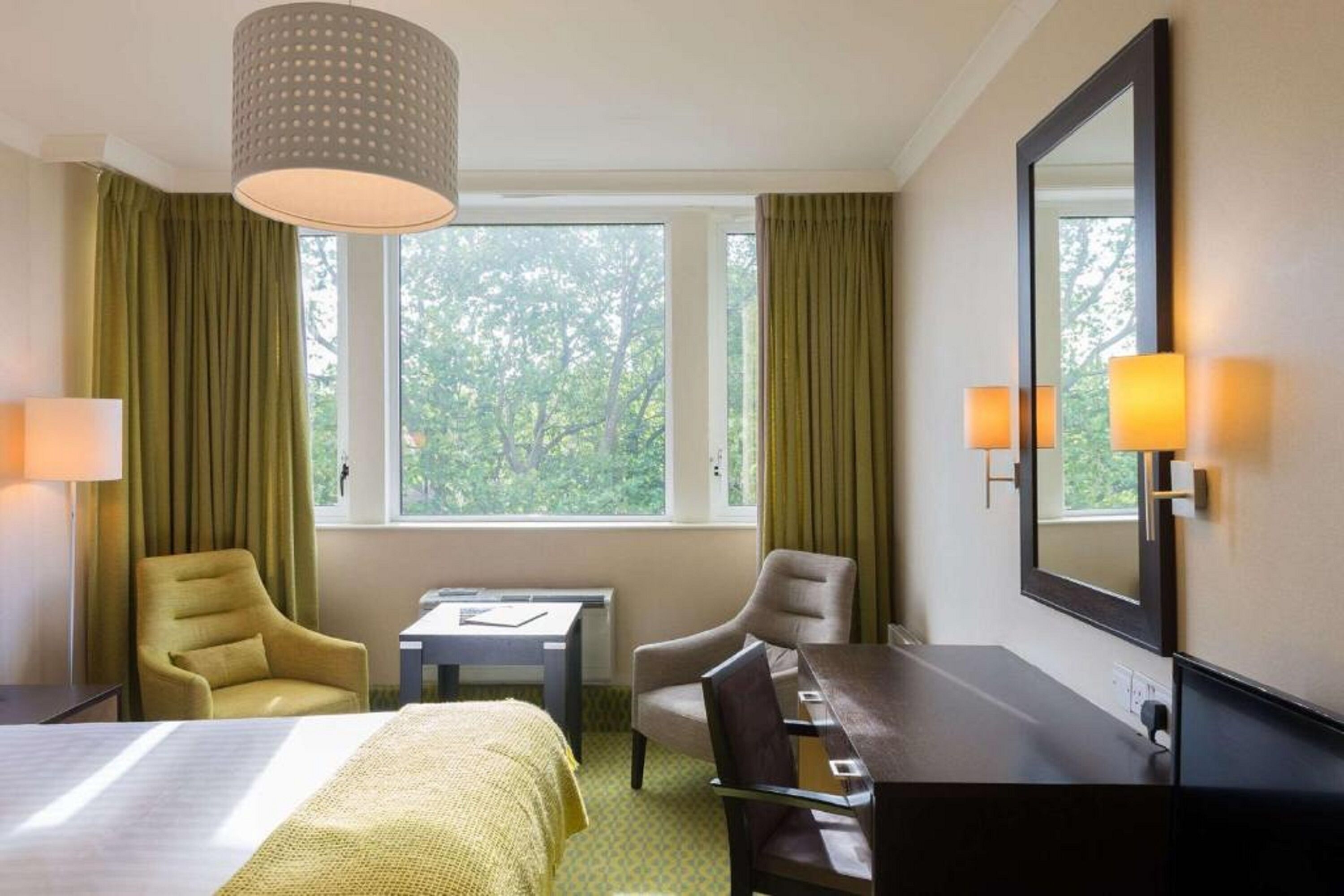 A hotel room with a bed, two armchairs, a desk, a lamp, a mirror, and a window with curtains.