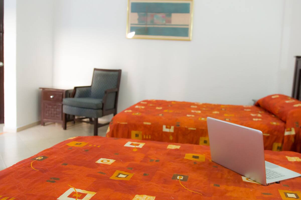 Een hotelkamer met twee bedden voorzien van oranje beddegoed met een patroon, een laptop op een van de bedden, een stoel en een kleine tafel.