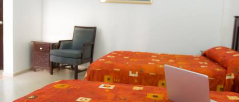 Habitación de hotel con dos camas cubiertas con colchas anaranjadas estampadas, una laptop sobre una cama, una silla y una mesita.