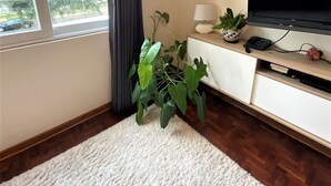 A living room with a large window, a flat-screen TV, a white sofa, a small table with a lamp, and a potted plant. - Private Modern Apartment in Pardo - Miraflores (Lima)