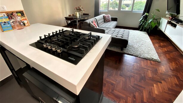 A modern kitchen with a central island, a dining area with a table and chairs, a sofa, and a large window with a view of buildings and greenery. - Private Modern Apartment in Pardo - Miraflores (Lima)
