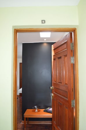 Una puerta de madera con un pestillo de metal, una pequeña mesa con un cuenco de metal y un libro, y una pared oscura al fondo.