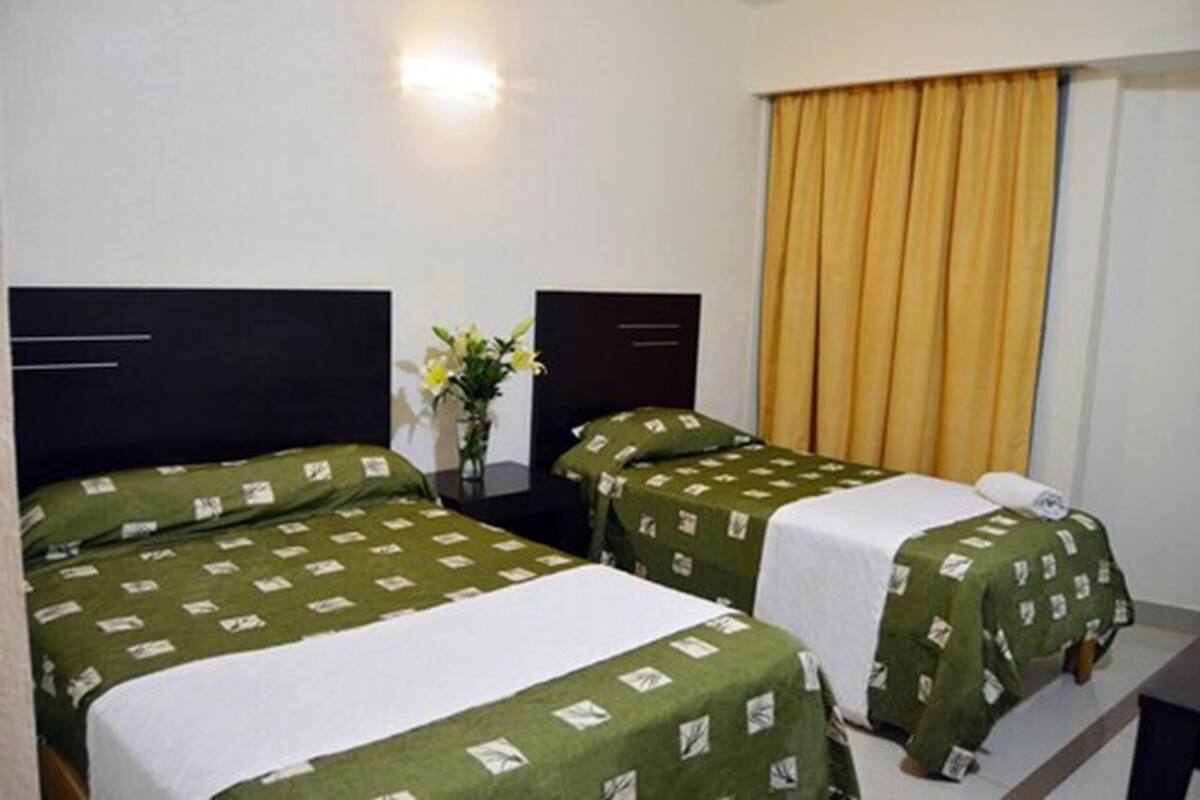 Habitación de hotel con dos camas individuales, una mesita de noche con un florero y una ventana con cortinas amarillas.