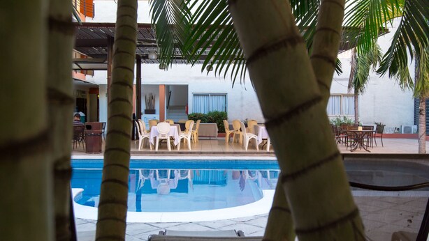 Zona de piscina con sillas y mesas, rodeada de palmeras y un edificio de fachada blanca.