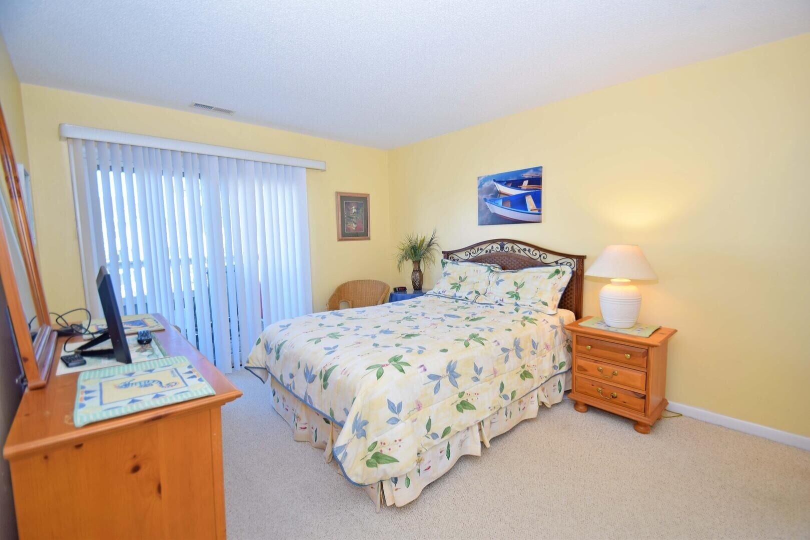 A bedroom with a bed, a desk, a chair, a nightstand, a lamp, and a window with blinds.