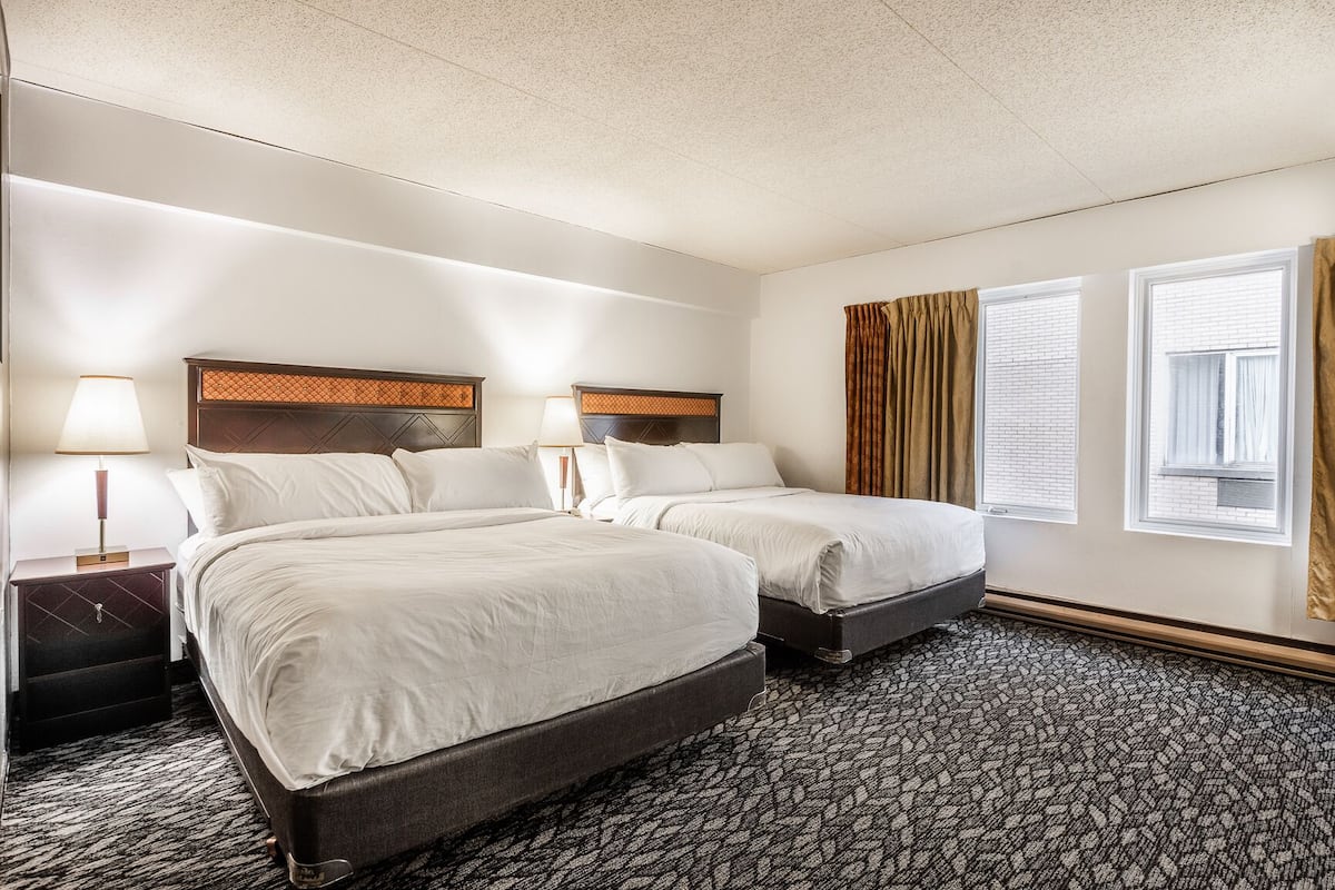 A hotel room with two beds, a nightstand with a lamp, a window with curtains, and a patterned carpet.