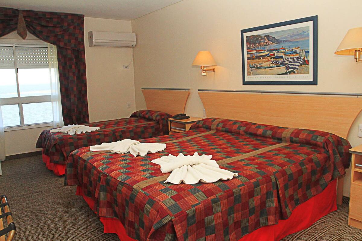 A hotel room with two beds, a painting of a coastal scene, and a window with curtains.